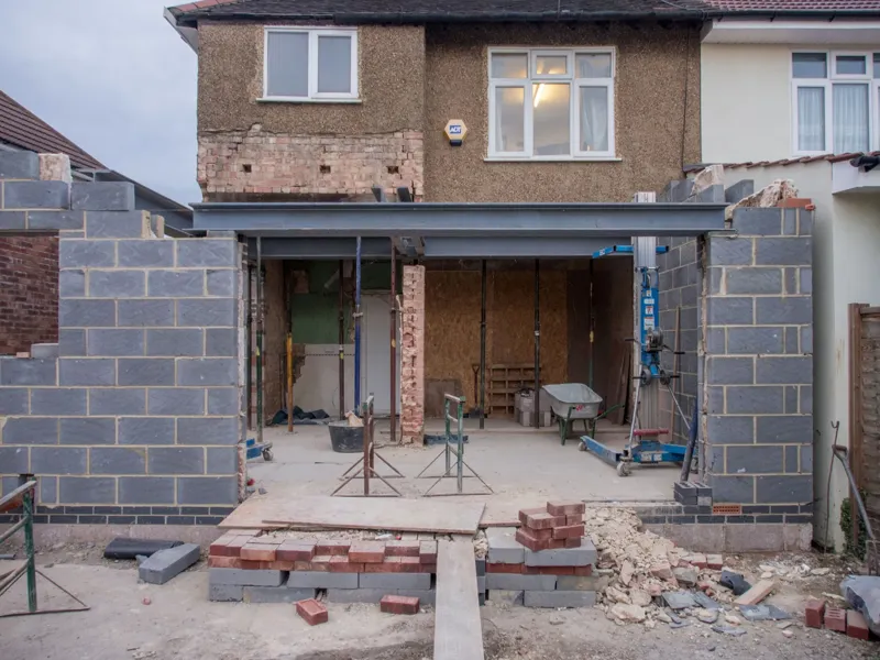 Brick house under construction with a large new opening in the rear wall spanned by a steel beam