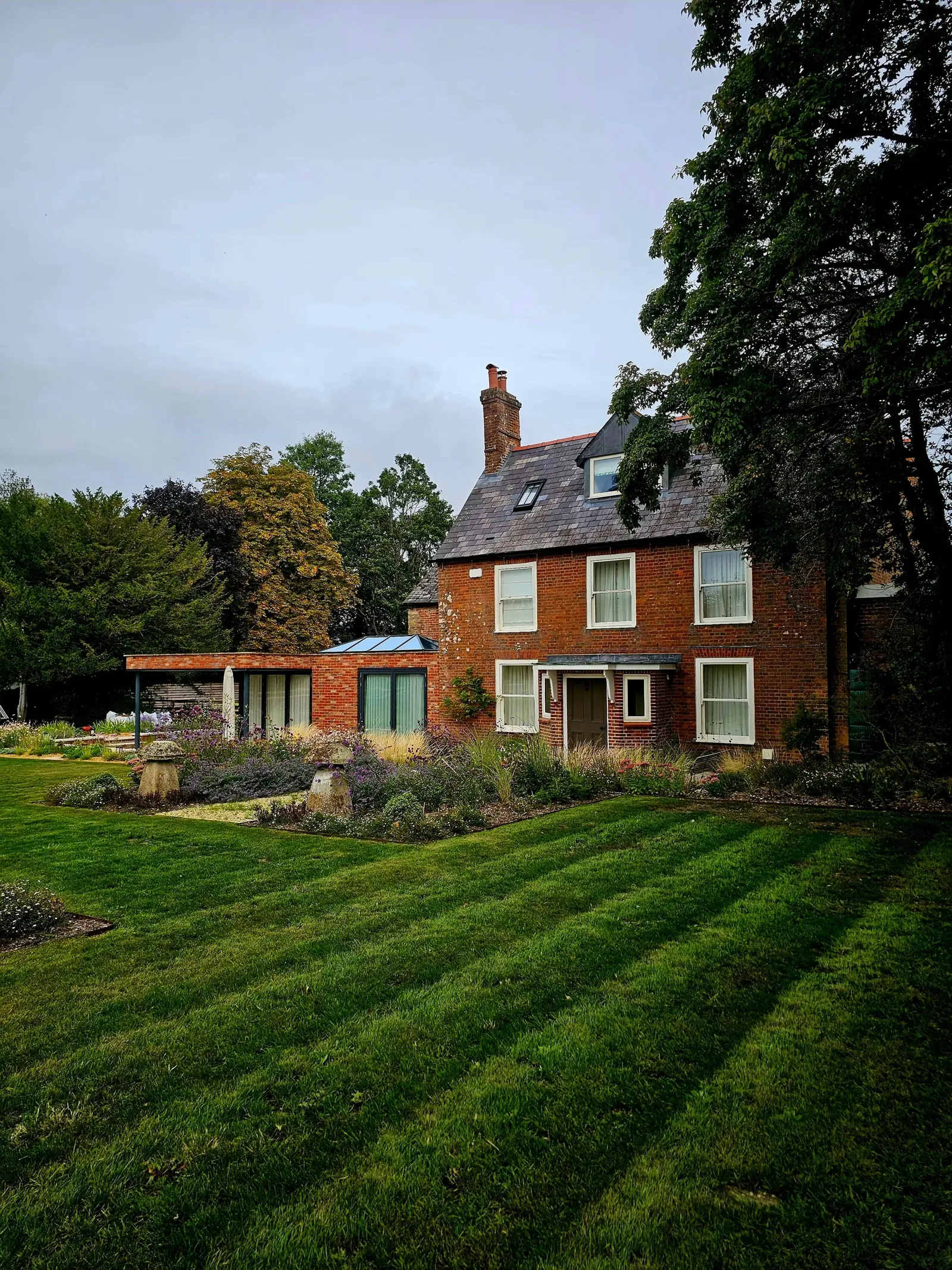Red-brick house with a slate roof and roof windows, a low glazed extension on its garden side and a striped lawn in front