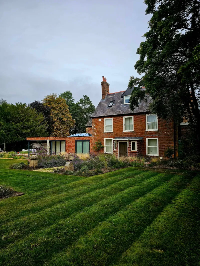 Red-brick house with a slate roof and roof windows, a low glazed extension on its garden side and a striped lawn in front