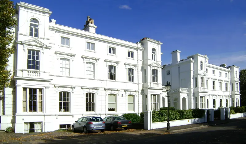 Row of tall Victorian brick houses with bay windows and white sash frames facing Richmond Green in south-west London
