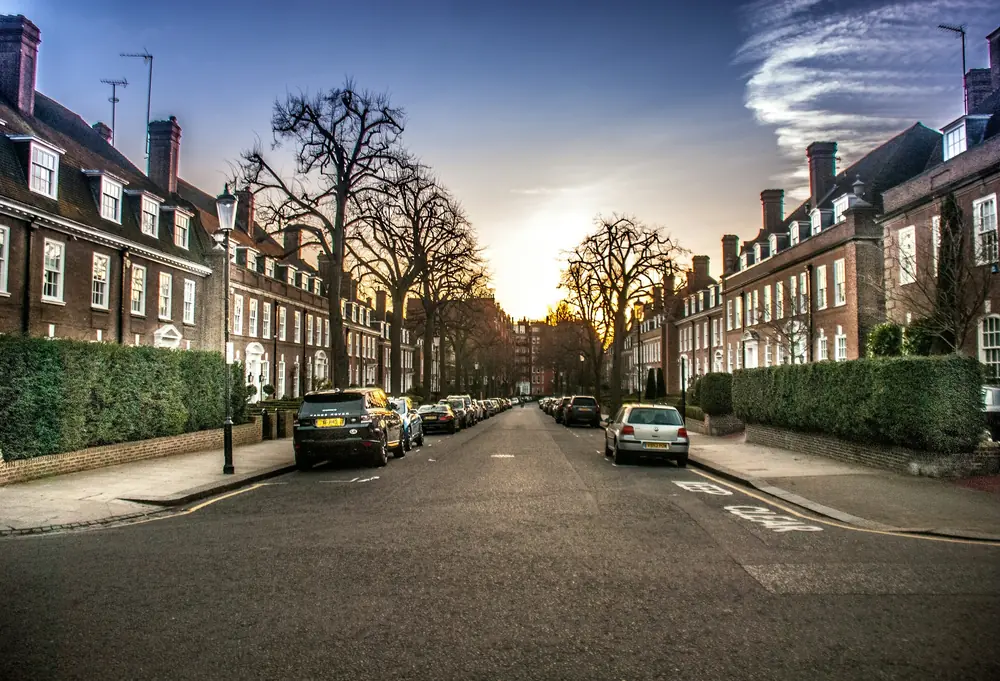 Tree-lined Kensington street of brick townhouses at sunset, streetlamps glowing under a dramatic dusk sky